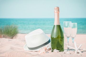Two glasses, a hat and a bottle of champagne on a sandy beach with seashells and green grass on a blue sea background