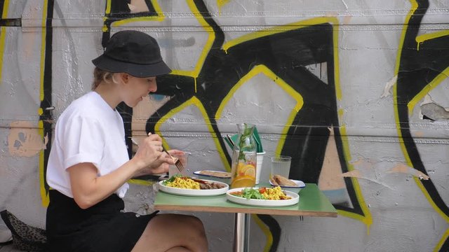 Side view of a cute girl in a bucket hat and white t-shirt eating her meal sitting next to graffiti wall in urban cafe. 