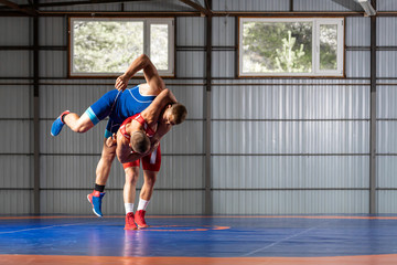 Two young men in blue and red wrestling tights are wrestlng and making a thigh throw, wrestling on a blue wrestling carpet in the gym. The concept of fair wrestling