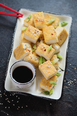 White plate with pan fried tofu cheese, green onion, sesame and soy sauce over dark brown stone surface, vertical shot