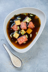 Miso soup with salmon, tofu cheese, wakame, sesame and green onion in a grey bowl, vertical shot on a light-blue stone surface