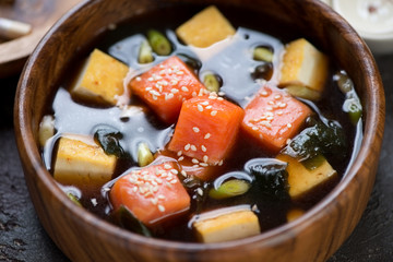 Close-up of miso soup with salmon fillet, chunks of tofu cheese, green onion and wakame served in a wooden bowl, selective focus