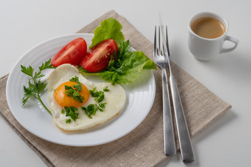 Healthy and hearty breakfast. Close-up of a fried eggs, tomato, herbs and espresso coffee. Served on a white table with a linen napkin and cutlery.