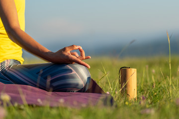 Caucasian woman sitting on her yoga mat holding her fingers in a mudra posture practicing meditation in nature with a koshi chime musical instrument sitting next to her body during a sunny morning.