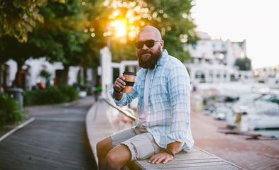 Portrait of young bearded man sitting at sunrise with cup of take away coffee, he using reusable...