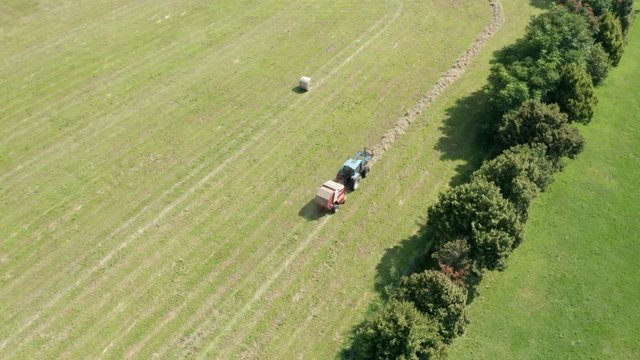 Aerial view of blue tractor with round baler discharging a fresh hay bale during harvesting