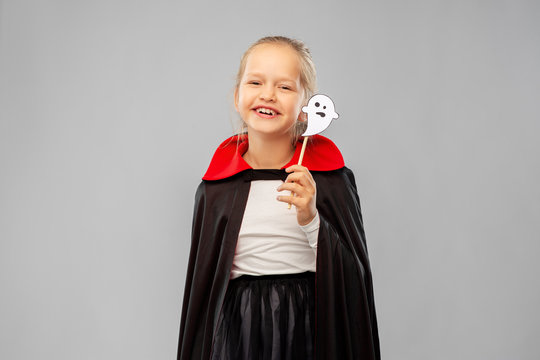 halloween party, holiday and childhood concept - happy smiling girl in black dracula costume with cape and ghost over grey background