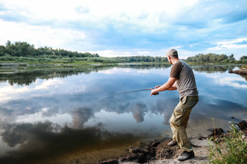 Fototapeta na wymiar Caucasian adult man fishing in evening with beautiful reflection sky and clouds on the river.