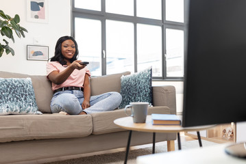 Obraz na płótnie Canvas people and leisure concept - happy smiling african american young woman with remote control watching tv at home