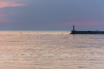 Fototapeta premium A lighthouse against the sunset sky. Beautiful peaceful seascape. Small ships on the sea horizon. Purple clouds and soft pink water. Seaport. Gulls in the sunset sky. Atmospheric evening landscape.
