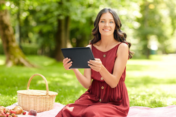 Fototapeta premium leisure, technology and people concept - happy smiling woman with tablet computer, picnic basket and food sitting on blanket at summer park
