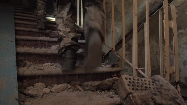 Close-up of fighters' legs in army boots climbing up stairs in abandoned ruined building during hostage rescue operation. Team of military men in camouflage storming upper floors of captured building