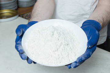A person holds a plate of flour in his hands dressed in blue gloves in his home kitchen. Steps to make bread at home.