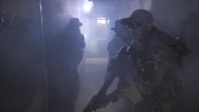 Small group of fully equipped soldiers alertly frozen in smoky corridor suspecting presence of enemy. One fighter with weapon at ready running in doorway while another covering comrade by shooting
