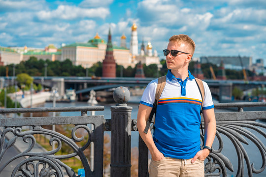 A young man with blond hair and sunglasses stands on a bridge overlooking the Kremlin and the river in Moscow