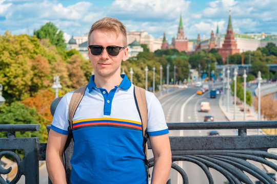 A young man with blond hair and sunglasses stands on a bridge overlooking the Kremlin and the river in Moscow