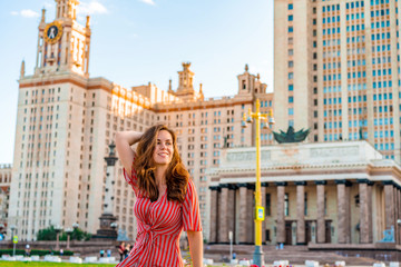Obraz premium Cute young woman in a red dress poses against the background of the main University of Russia, Moscow high-rise