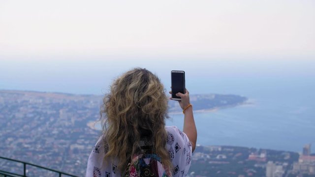 Back view of woman holding smartphone and taking photos of beautiful seascape. Rear view of attractive young woman standing on observation point and photographing coastal town and sea bay