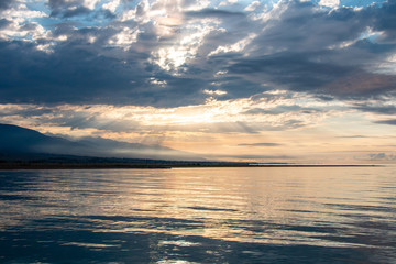 Naklejka na ściany i meble Beautiful sun rays passing through the clouds above the calm lake with reflection on it and with mountain silhouettes on background. Morning mood. Blue cloudy sky..