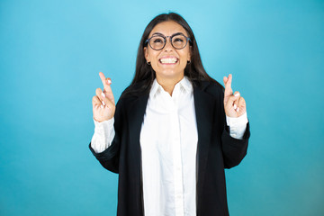 Young beautiful business woman over isolated blue background gesturing finger crossed smiling with...