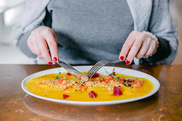 Woman in a grey jumper is eating a beautifully decorated pumpkin dish decorated with red beetroot cubes, greens and seeds. Close-up of woman's hands with fork and a knife. In the restaurant.