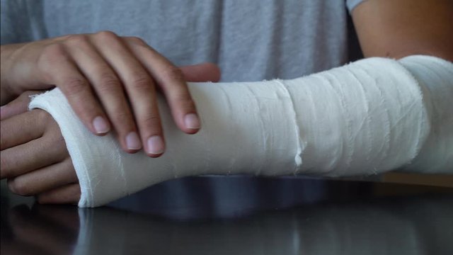 Man doing self massage on broken arm wrapped in plaster cast. Closeup of patient rubbing and stroking disabled limb while sitting at table. Concept of healthcare