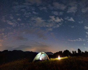 Illuminated tent and campfire under incredible starry sky, summer evening in the mountains, beauty of the nature. Night camping