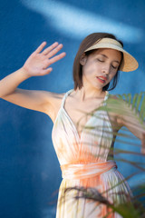 Woman in batik dress stands near of green plants at big blue wall, with light and shadow.