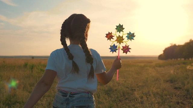 A happy, sweet girl runs across a field at sunset with a windmill in her hand. A child plays in the open air on a meadow.