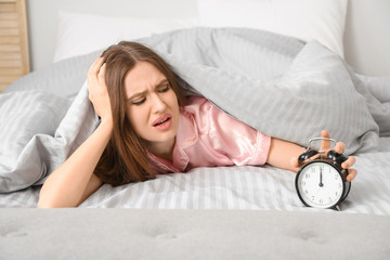 Young woman with alarm clock suffering from insomnia in bedroom