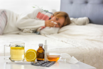 Selective focus on alternative medicine on the night table (hot drink, lemon, honey, pills, thermometer, nasal spray and crumpled handkerchiefs. Young sick woman laying on bed