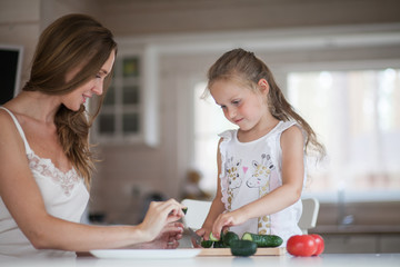 Naklejka premium Happy family mother and child posing at home. Beautiful young mom and little daughter having fun and preparing vegetables for salad in a white kitchen in a Scandinavian style interior. Healthy food.