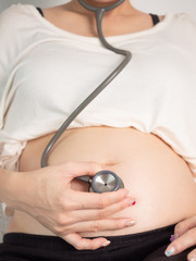 Pregnant Asian woman using stethoscope listening to baby heartbeat in white bedroom. 