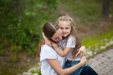 Fototapeta premium Happy smiling mother and her cute emotional little daughter child in white shirts and denim jeans are hugging and having fun outdoor in nature at countryside. Spending summer time with family.