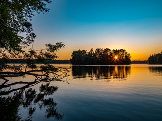 Sunset over Pymatuning Lake in Pennsylvania at the end of summer.  A fallen tree in the foreground and the sunburst coming through the trees on an island in the background and the sky orange and blue.
