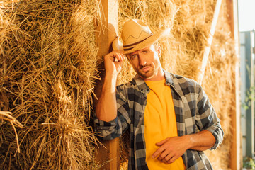 rancher in plaid shirt looking at camera and touching straw hat while leaning on hay stack