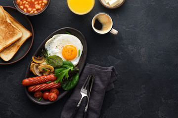 Traditional full English breakfast with fried eggs, sausages, beans, mushrooms, grilled tomatoes on black background.
