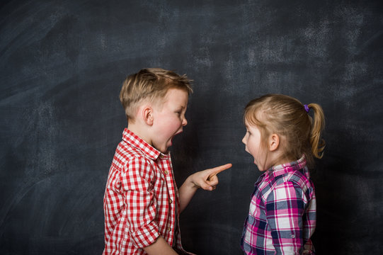Boy and girl pointing fingers to each other. Raging kids children shouting to each other against blackboard background. blame