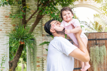 Fototapeta premium Cheerful father holding and kiss baby daughter in his hand while relax time at home. Mixed race little lovely girl with daddy carrying in the garden . Happy father day concept.