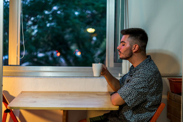 Joven con taza de cafe trabajando en el ordenador junto a la ventana