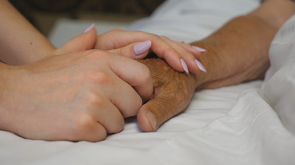Granddaughter holds and comforts hand of her grandmother in medical clinic. Daughter gently touches wrinkled arm of sick mature mother lying in bed hospital. Concept of care and love. Slow motion