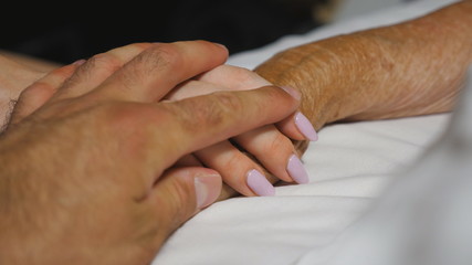 Fototapeta premium Granddaughter and grandson gently touch hand of their grandma lying in bed hospital. Young woman and man comfort wrinkled arm of sick grandmother in medical clinic. Concept of care and love. Close up