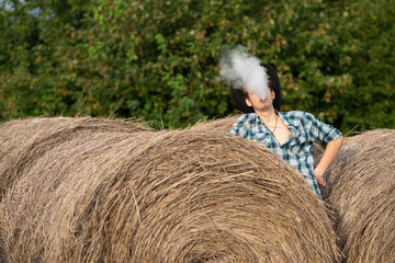 Naklejka na ściany i meble Vape man. Portrait of a handsome young white guy in a cowboy hat vaping an electronic cigarette near the haystack. Countryside lifestyle concept.