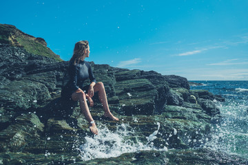 Young woman sitting on rocks by sea with waves crashing in