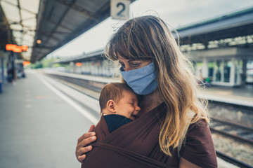 Fototapeta na wymiar Young mother with baby in sling wearing face mask at train station