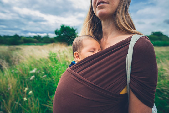 Young mother with baby in sling standing outdoors in meadow