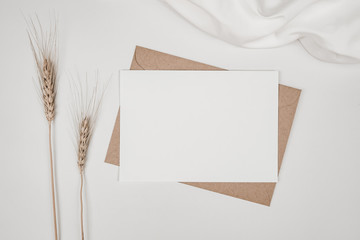 Blank white paper on brown paper envelope with Barley dry flower and white cloth. Mock-up of horizontal blank greeting card. Top view of Craft envelope on white background. Flat lay minimalism