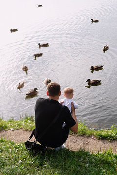 man with small beren in his arms is sitting on  river bank, ducks swim on  river