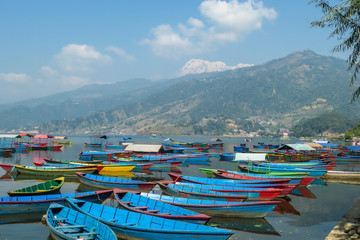 A view on Phewa Lake in Pokhara, Nepal with many colorful boats anchored around the shore. There are high Himalayan ranges in the back. Calm surface of the lake. Clear and sunny day. Serenity
