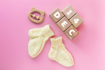 14 DECEMBER text on wood cubes, pair of cute knitted baby socks and wooden teether bear on pink background. Date of child birth. Newborn concept. Flat lay, top view with copy space.